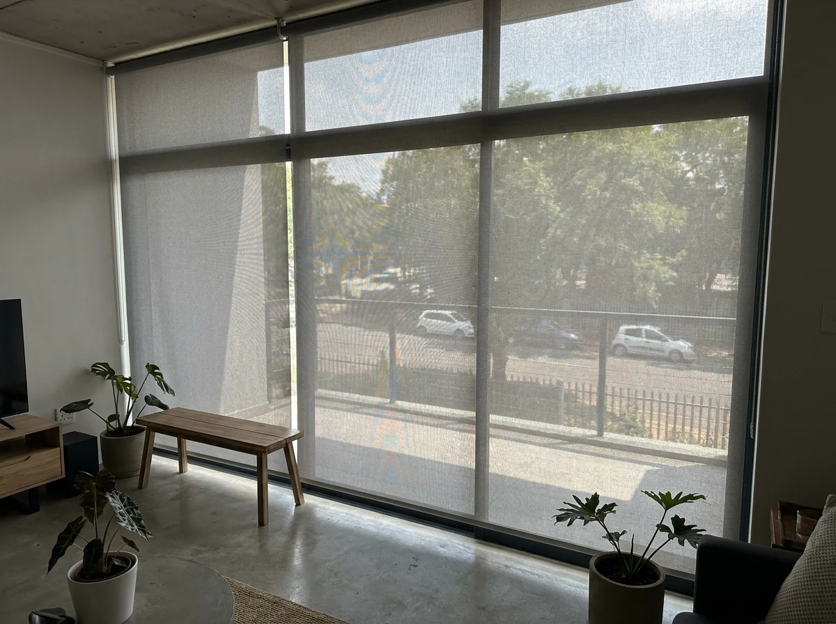 Sunscreen mesh roller blind lowered over a large window in a modern Brooklyn apartment, tree-lined street faintly visible through the weave