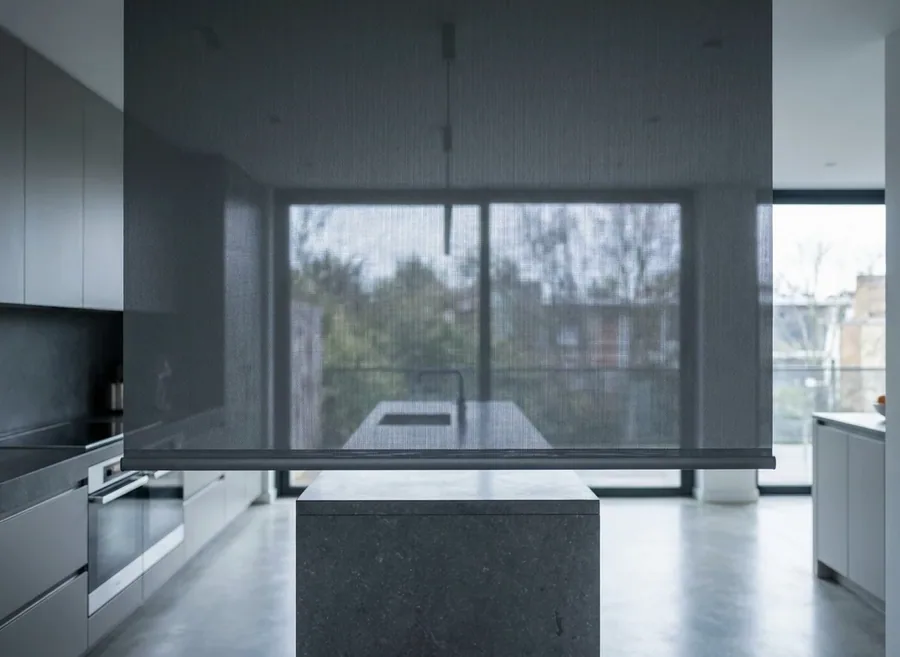 Sunscreen roller blind in mesh fabric lowered over a wide window, the garden still visible through the weave
